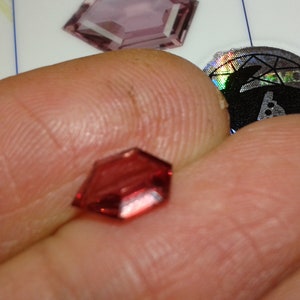 Puede incluir: Una gema roja facetada sostenida entre los dedos. La gema tiene forma hexagonal con múltiples facetas, reflejando la luz. Otras gemas y una pegatina con un diseño de conejo están en el fondo.