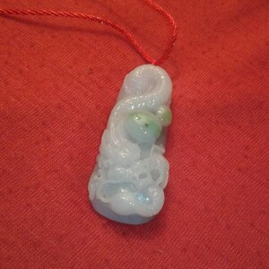 Puede incluir: Un colgante de jade tallado en tonos azul claro y verde, suspendido de un cordón rojo. El colgante presenta un diseño orgánico detallado. El fondo es una tela roja lisa.