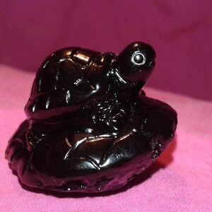 Puede incluir: Una figura de tortuga negra brillante. La tortuga está esculpida con detalles, incluyendo un caparazón texturizado y una pequeña cabeza con ojos visibles. La figura se asienta sobre una base texturizada, sobre un fondo rosa.