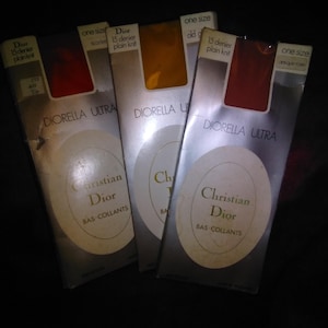 Op de afbeelding: Drie verpakkingen Diorella Ultra kousen in verschillende kleuren, waaronder rood en goud. Elke verpakking toont het Christian Dior logo en de tekst "BAS-COLLANTS". De verpakkingen zijn gemarkeerd met "one size" en "15 denier plain knit".
