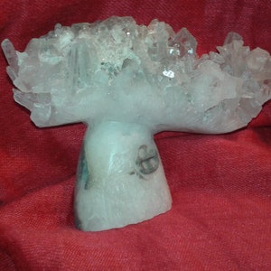 May include: A clear, crystalline quartz cluster sculpture. The quartz has a mushroom-like shape, with a rounded top and a stem. The sculpture is set against a red background.