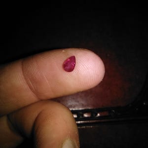 Könnte beinhalten: Ein kleiner, birnenförmiger, tiefroter Edelstein, möglicherweise ein Rubin, liegt auf einer Fingerspitze. Der Edelstein ist glatt und poliert und reflektiert das Licht. Der Hintergrund ist dunkel und betont die Farbe und Form des Edelsteins. Das Bild ist eine Nahaufnahme.