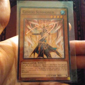 以下が含まれることがあります： 「ジェミニ・サモナー」をフィーチャーした遊戯王トレーディングカード。カードは金とオレンジ色の背景で、黒と青のローブを着たキャラクターがいます。カードテキストには「Spellcaster/Effect」と「ATK/1500 DEF/ 0」が含まれています。