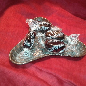 May include: A small, ornate silver dish holds several individually wrapped candies. The candies are dark brown with white text that reads "Hakemans". The dish sits on a red fabric surface.