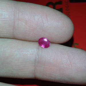 Könnte beinhalten: Ein kleiner, oval geformter, leuchtend rosa Rubin-Edelstein. Der Edelstein wird zwischen zwei Fingern gehalten, wodurch seine polierte Oberfläche und seine leuchtende Farbe zur Geltung kommen. Ein Rubin ist ein Edelstein, der oft in Schmuck verwendet wird.