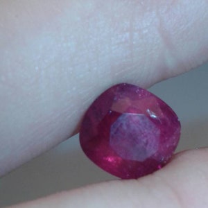 May include: A faceted, cushion-cut ruby gemstone. The gem is a deep, saturated red color with internal reflections. The stone is held between two fingers, showcasing its polished surfaces and clarity.