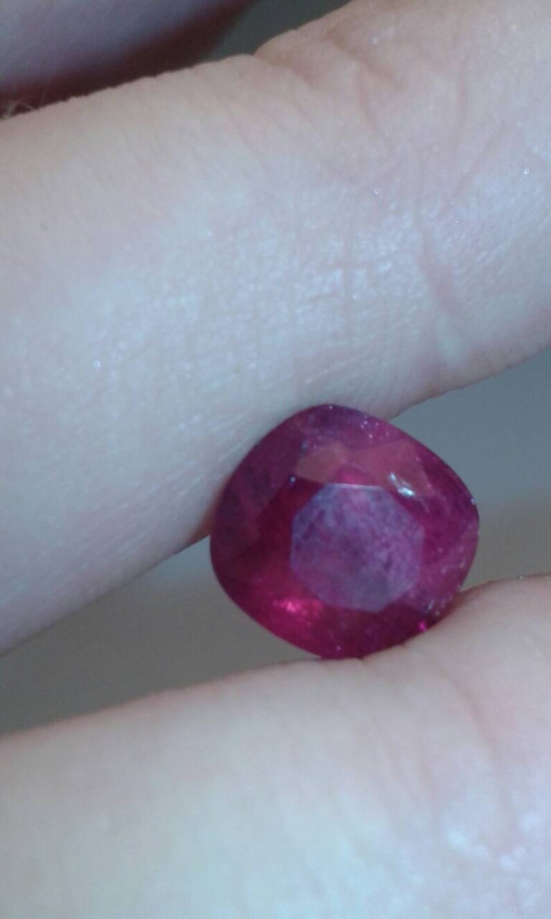 May include: A faceted, cushion-cut ruby gemstone. The gem is a deep, saturated red color with internal reflections. The stone is held between two fingers, showcasing its polished surfaces and clarity.