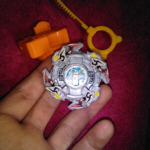 May include: A silver and blue spinning top toy with red, yellow, and black accents. The toy is held in a hand, with an orange launcher and a yellow ripcord nearby. The toy has a detailed design and is likely a collectible.
