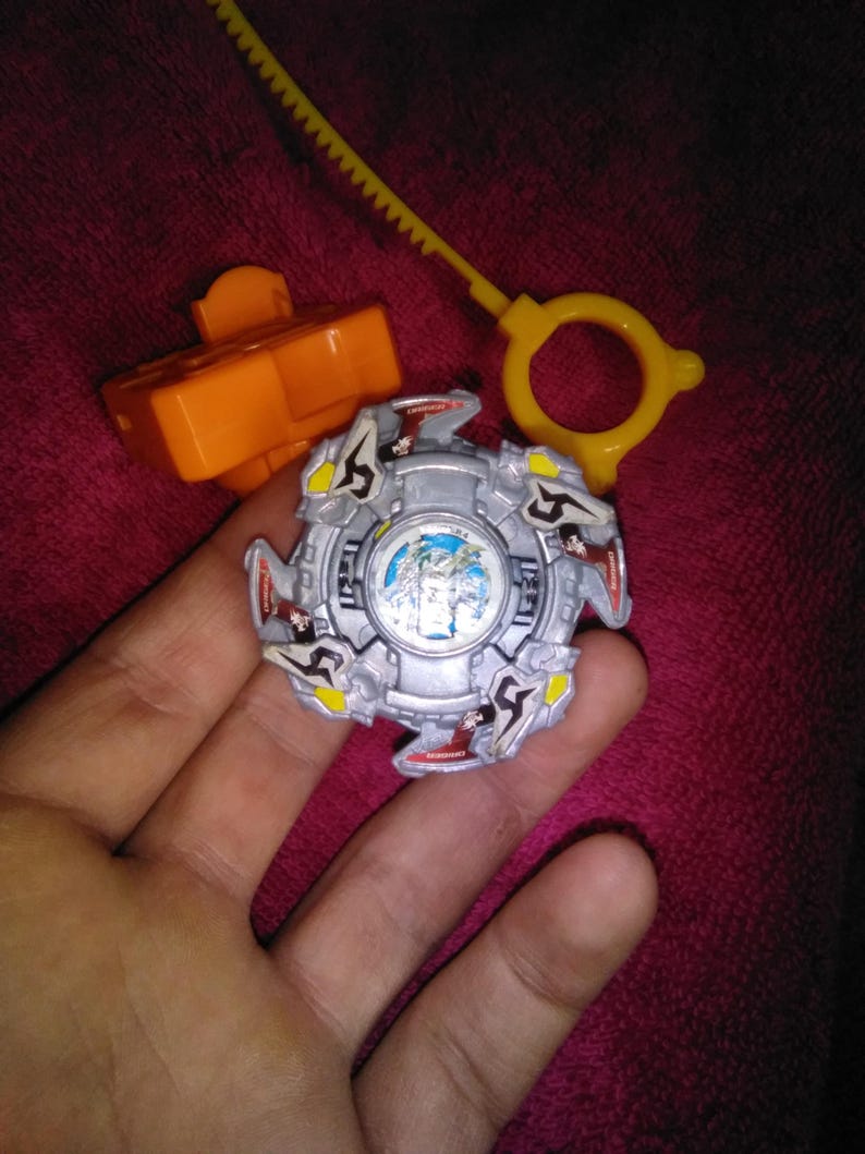 May include: A silver and blue spinning top toy with red, yellow, and black accents. The toy is held in a hand, with an orange launcher and a yellow ripcord nearby. The toy has a detailed design and is likely a collectible.