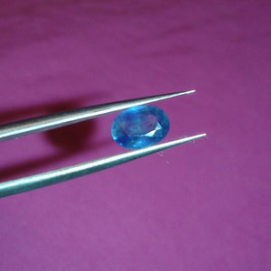 May include: A brilliant blue oval gemstone is held by silver tweezers against a purple background. The gemstone has facets that reflect light, creating a sparkling effect. The tweezers are positioned to display the gem.
