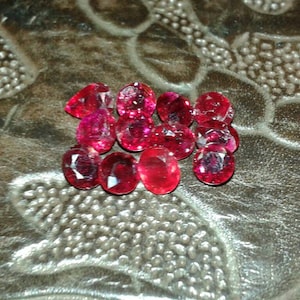May include: A collection of faceted, oval and round-cut, deep red gemstones. The gems are scattered on a textured, brown surface. The stones have a brilliant, reflective quality, suggesting they are high-quality rubies or similar precious stones.