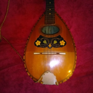 Peut inclure: Mandoline vintage avec un corps en forme de poire et une finition en bois. L'instrument présente des motifs floraux décoratifs jaunes et noirs, avec une rosace ovale bleue. Une corde est attachée au corps.