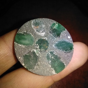 May include: A circular, grey stone featuring embedded green gemstones. The gemstones are irregularly shaped and of varying sizes. The stone is held in a person's hand, with the background out of focus.