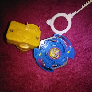 May include: A blue and silver spinning top toy with a yellow and purple center design, a yellow launcher, and a white pull string. The toy is designed for spinning and battling.