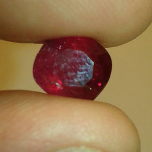 May include: A faceted, oval-shaped ruby gemstone, held between two fingers. The gem is a deep, saturated red color with internal reflections. The facets catch the light, creating a sparkling effect. The background is a soft, blurred neutral tone.