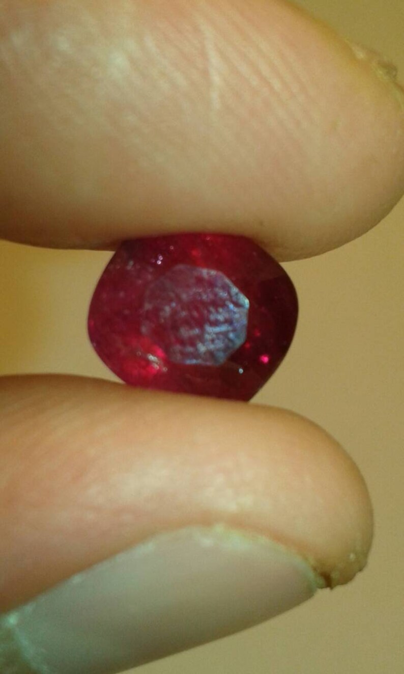 May include: A faceted, oval-shaped ruby gemstone, held between two fingers. The gem is a deep, saturated red color with internal reflections. The facets catch the light, creating a sparkling effect. The background is a soft, blurred neutral tone.