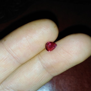 Könnte beinhalten: Ein kleiner, herzförmiger, facettierter, roter Edelstein. Der Edelstein wird zwischen zwei Fingern gehalten, was seine leuchtende Farbe und seinen Schliff zeigt. Der Hintergrund ist dunkel und betont den Glanz des Edelsteins.