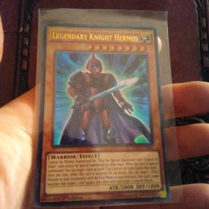 May include: A Legendary Knight Hermos trading card, featuring a warrior in red armor with a sword. The card has a blue and orange background and text that reads "WARRIOR/EFFECT" and "ATK/2800 DEF/1800".