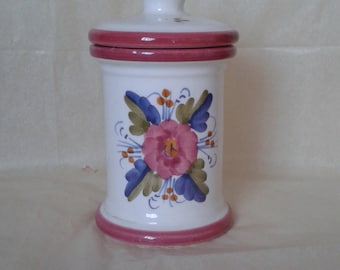 May include: White ceramic jar with a lid, featuring a floral design in pink, blue, and green. The jar has a cylindrical shape with a pink rim at the top and bottom. The lid is white with a small finial.