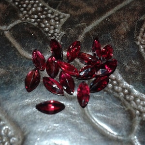 May include: A collection of marquise-cut, deep red gemstones. The gems are scattered on a textured, dark surface, catching the light and displaying a rich, glossy appearance. These faceted stones are ideal for jewelry making.