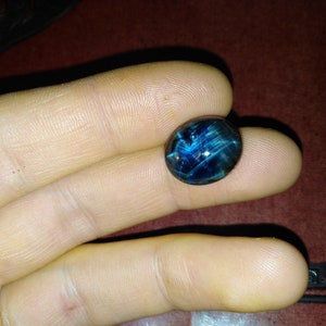 May include: A polished, oval-shaped gemstone with a deep blue hue and a star-like pattern. The gem's surface reflects light, creating a captivating visual effect. The stone is held between fingers, showcasing its size and detail.