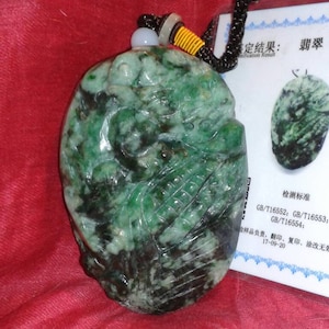 Può includere: Un ciondolo in giada scolpita con un motivo verde e nero, sospeso a un cordino nero. Il ciondolo presenta un design dettagliato ed è esposto su sfondo rosso. È visibile una scheda bianca con testo cinese.