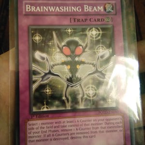 May include: A Yu-Gi-Oh! Brainwashing Beam trap card, 1st Edition, encased in a protective sleeve. The card features a dark alien-like creature with red eyes and a detailed background. The card's text describes its effect.