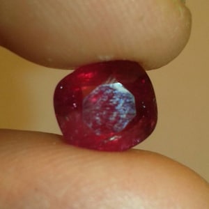 May include: A faceted, deep red gemstone, held between two fingers. The gem has a slightly rounded, square shape with internal reflections. The color is a rich, saturated red, with a hint of lighter tones within the facets.