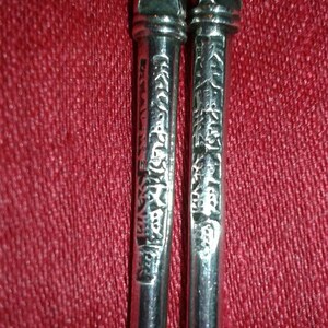 May include: Close-up of two silver chopsticks with ornate, geometric tops and engraved characters along the shafts. The chopsticks are set against a textured, red background.