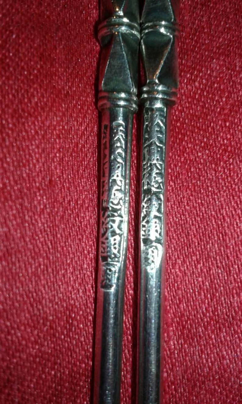 May include: Close-up of two silver chopsticks with ornate, geometric tops and engraved characters along the shafts. The chopsticks are set against a textured, red background.