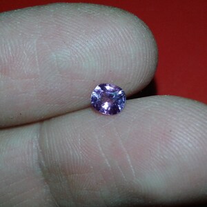 May include: A small, round, faceted gemstone with a light purple hue. The gem is held between two fingers against a red background. The gem's facets catch the light, creating a sparkling effect.