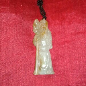 Op de afbeelding: Een gebeeldhouwde jade hanger met een geklede figuur die een staf vasthoudt. De hanger is lichtgroen met bruine accenten en hangt aan een zwart koord. De achtergrond is een rode stof met textuur.