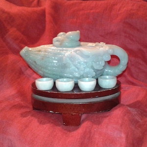 Könnte beinhalten: Ein geschnitztes Jade-Teeservice mit einer Teekanne mit Drachendesign und vier kleinen Tassen. Das Set steht auf einem Holzständer vor rotem Hintergrund. Teekanne und Tassen sind hellgrün.