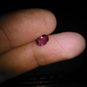 May include: A small, oval-shaped, pink gemstone is held between two fingers. The gem has a polished surface and a deep, saturated color. The background is dark, emphasizing the gem's brilliance. This could be a ruby or pink sapphire.