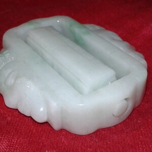 Può includere: Una pietra di giada scolpita, di colore bianco e verde chiaro. Il design rettangolare presenta tre sezioni rialzate e un piccolo foro. La pietra è presentata su uno sfondo rosso, evidenziando i suoi dettagli.