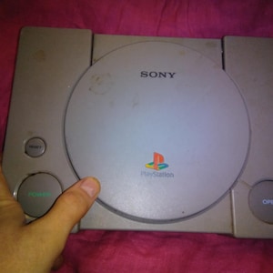 Peut inclure: Une console PlayStation Sony grise. La console a un lecteur de disque circulaire, le logo PlayStation et le mot "SONY" imprimé sur le dessus. Elle comporte des boutons pour l'alimentation, la réinitialisation et l'ouverture. La console est tenue par une main.