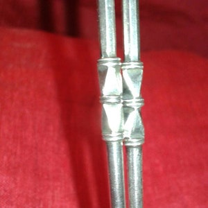 May include: Close-up of two silver metal straws with decorative geometric accents. The straws are set against a red background, highlighting their metallic sheen. The design features a series of connected, diamond-shaped elements.