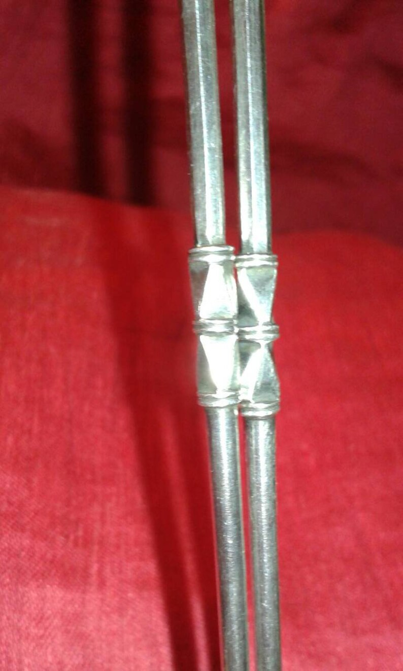 May include: Close-up of two silver metal straws with decorative geometric accents. The straws are set against a red background, highlighting their metallic sheen. The design features a series of connected, diamond-shaped elements.