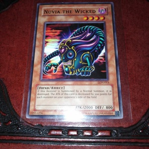 Puede incluir: Una carta coleccionable de Yu-Gi-Oh! con "Nuvia the Wicked". La carta tiene una ilustración de un monstruo turquesa y morado con detalles dorados. La carta está en una funda protectora y tiene texto, incluyendo "ATK/2000 DEF/800".