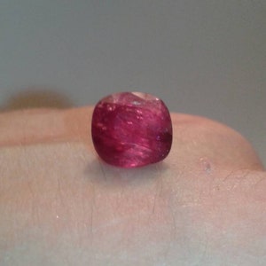May include: A cushion-cut, translucent, deep red gemstone. The gem is set against a neutral background, showcasing its color and clarity. The stone's facets catch the light, creating a sparkling effect. This gemstone is ideal for jewelry.