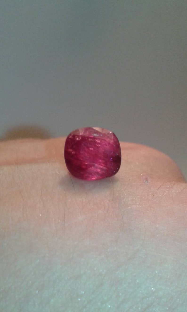 May include: A cushion-cut, translucent, deep red gemstone. The gem is set against a neutral background, showcasing its color and clarity. The stone's facets catch the light, creating a sparkling effect. This gemstone is ideal for jewelry.