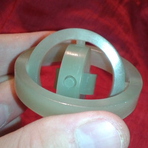Puede incluir: Un juguete de rompecabezas verde pálido con un anillo exterior circular y un mecanismo interno. El juguete se sostiene sobre un fondo rojo. El diseño presenta formas redondeadas e intersecantes.