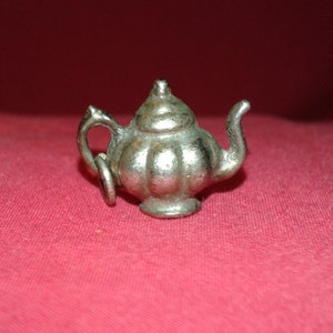 Peut inclure: Un petit charme en forme de théière argentée, avec un design détaillé. La théière a un bec courbé, une anse et un couvercle avec un petit bouton. Elle est posée sur un fond rouge, mettant en valeur sa finition métallique et ses détails.