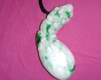 Puede incluir: Colgante de jade blanco y verde con un cordón negro. El jade está tallado en una forma curva con vetas y manchas verdes. El colgante cuelga sobre un fondo rosa.