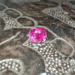 May include: A vibrant, cushion-cut, pink gemstone. The gem is set against a dark, textured surface with a repeating pattern. The gemstone's facets reflect light, creating a sparkling effect. The overall composition highlights the gem's color and shape.