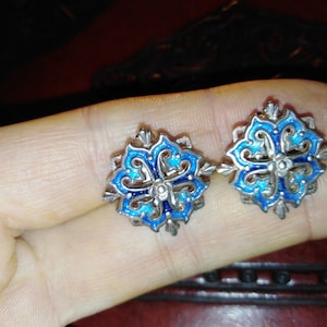 May include: A pair of ornate silver-toned earrings with a floral design. Each earring features a central blue enamel flower surrounded by a detailed silver filigree. The earrings are a vintage style.