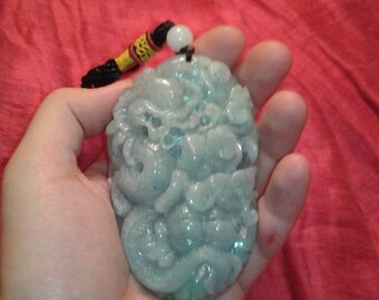 Puede incluir: Un colgante de jade verde claro con intrincadas tallas, sostenido en una mano sobre un fondo rojo. El colgante está sujeto a un cordón negro y amarillo con una cuenta blanca. El diseño es detallado.