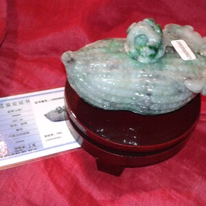 May include: A carved jade sculpture of a gourd with a small flower on top, displayed on a dark wooden stand. The jade is primarily light green with some darker green accents. A certificate is visible next to the sculpture.