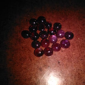 Puede incluir: Un conjunto de canicas de color rojo oscuro y púrpura. Las canicas tienen un acabado brillante y varían en tonos de rojo y púrpura. Las canicas están dispersas sobre una superficie marrón.