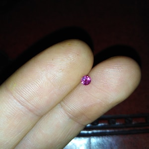 May include: A small, round, vibrant pink gemstone, likely a ruby or pink sapphire, rests on a person's fingers. The gem is a deep, saturated pink color and appears to be faceted. The background is a dark, blurred surface.